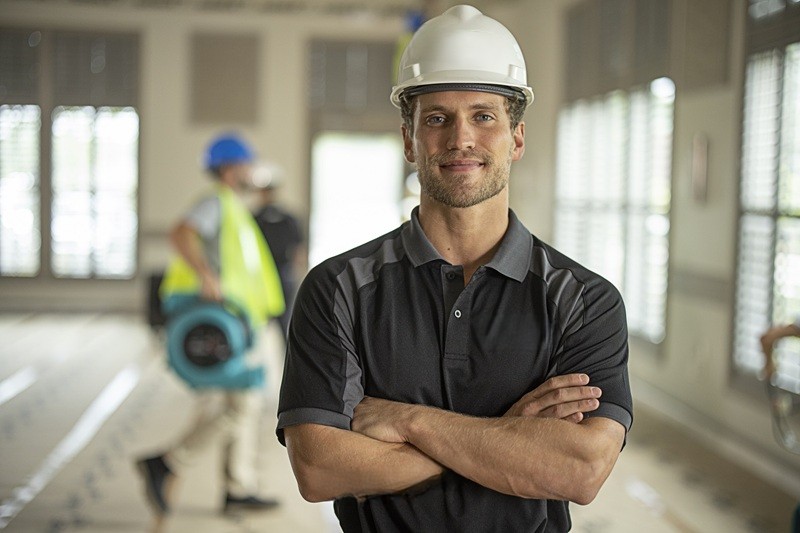 SERVICEMASTER EMERGENCY RESTORATION EXPERTS EMPLOYMENT A SERVICEMASTER EMERGENCY RESTORATION EXPERTS TECHINICIAN POSES CONFIDENTLY FOR THE CAMERA. ANOTHER TECHNICIAN IS IN THE BACKGROUND MOVING DRYING EQUIPMENT INTO THE COMMERCIAL PROPERTY
