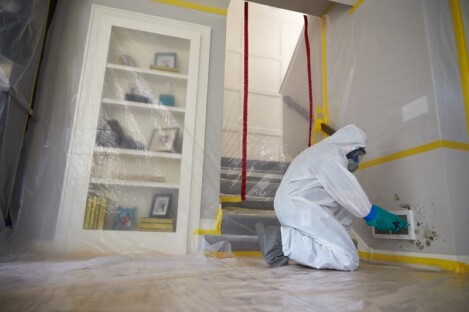 A RESIDENTIAL MOLD REMEDIATION TECHNICIAN IS DRESSED IN PROTECTIVE COVERALLS AND A RESPIRATOR AS HE BEGINS TO UNSCREW A REGISTER COVER FROM THE WALL TO BEGIN MOLD REMOVAL 